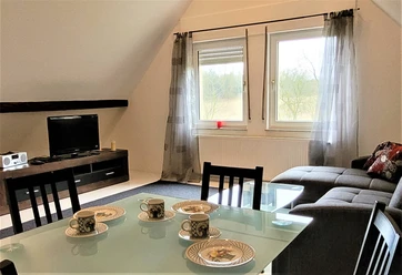FeWo Obereickum-Wohnzimmer - B Schlaupitz (15).jpg Wohnzimmer mit Ecksofa, Glastisch und Fernseher; Fenster bieten Aussicht auf grüne Landschaft.