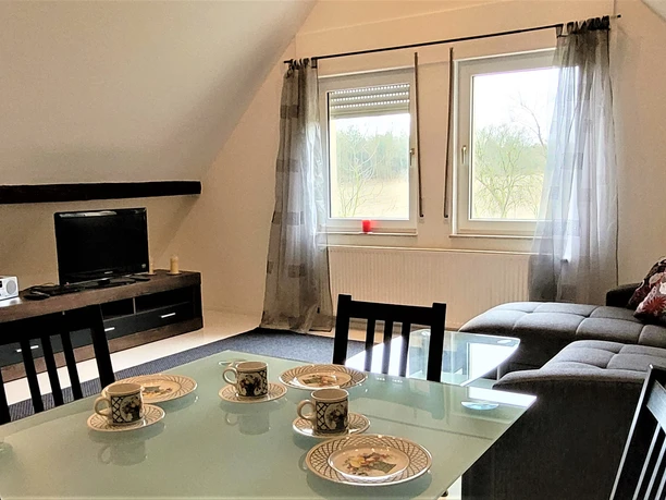 FeWo Obereickum-Wohnzimmer - B Schlaupitz (15).jpg Wohnzimmer mit Ecksofa, Glastisch und Fernseher; Fenster bieten Aussicht auf grüne Landschaft.