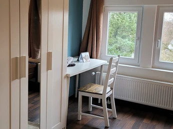 Helles Zimmer mit Schreibtisch, Stuhl und zwei Fenstern, die Blicke ins Grüne erlauben.