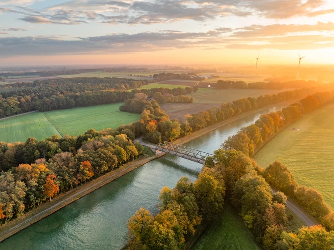 Mittellandkanal bei Bad Essen Luftaufnahme eines Kanals mit Brücke, umgeben von herbstlich gefärbten Bäumen bei Sonnenuntergang.Aerial view of a canal with a bridge, surrounded by autumn-colored trees at sunset.Luftfoto af en kanal med en bro, omgivet af efterårsfarvede træer ved solnedgang.Luchtfoto van een kanaal met een brug, omringd door herfstkleurige bomen bij zonsondergang.