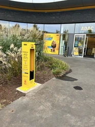 Gelber ADAC-Fahrradreparaturstation vor einem ADAC-Gebäude, umgeben von Ziergräsern und Pflanzen.