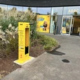 Gelber ADAC-Fahrradreparaturstation vor einem ADAC-Gebäude, umgeben von Ziergräsern und Pflanzen.