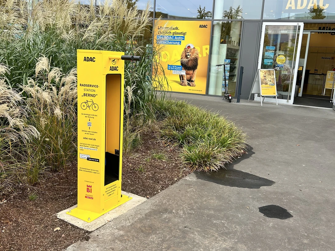 bielefeld-radservicestation-teutoburgerwald-bielefeld-marketing-adac owl Gelber ADAC-Fahrradreparaturstation vor einem ADAC-Gebäude, umgeben von Ziergräsern und Pflanzen.
