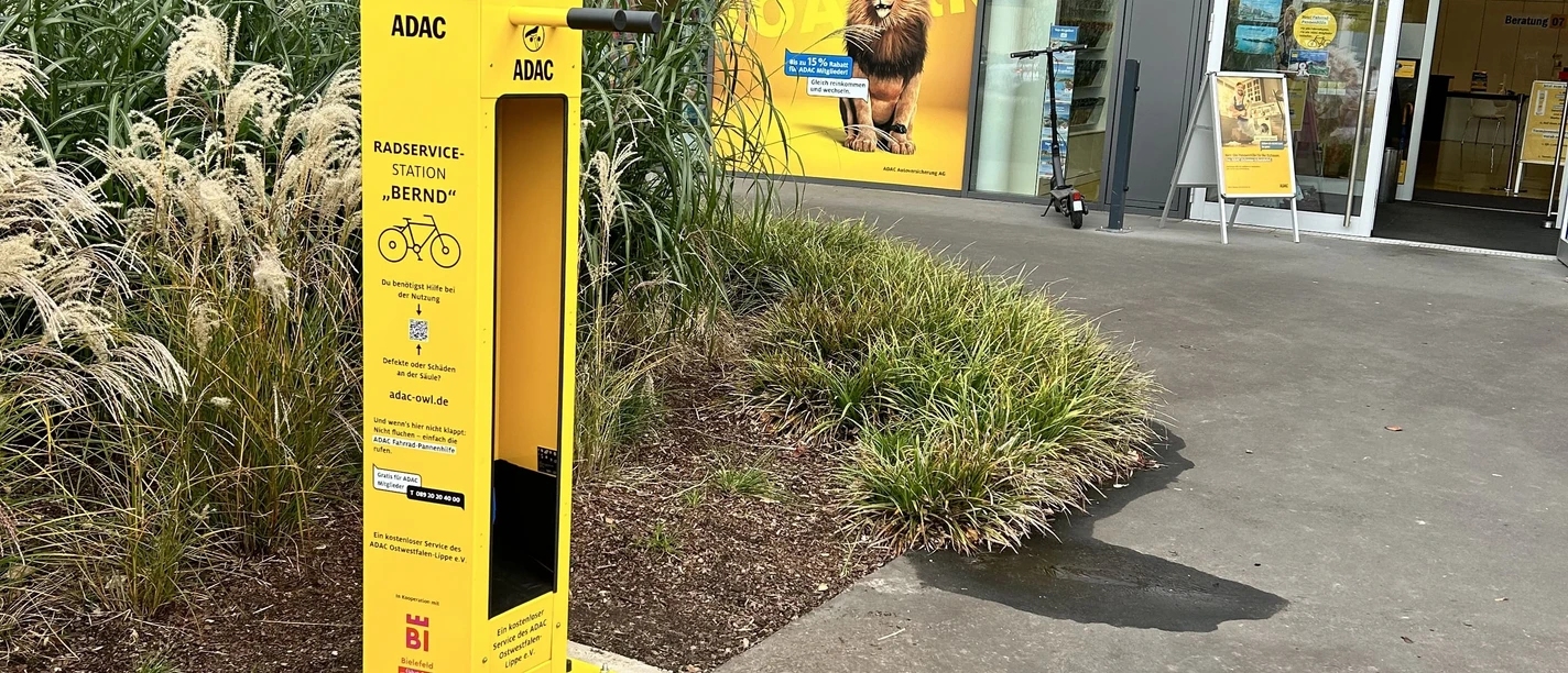 bielefeld-radservicestation-teutoburgerwald-bielefeld-marketing-adac owl Gelber ADAC-Fahrradreparaturstation vor einem ADAC-Gebäude, umgeben von Ziergräsern und Pflanzen.