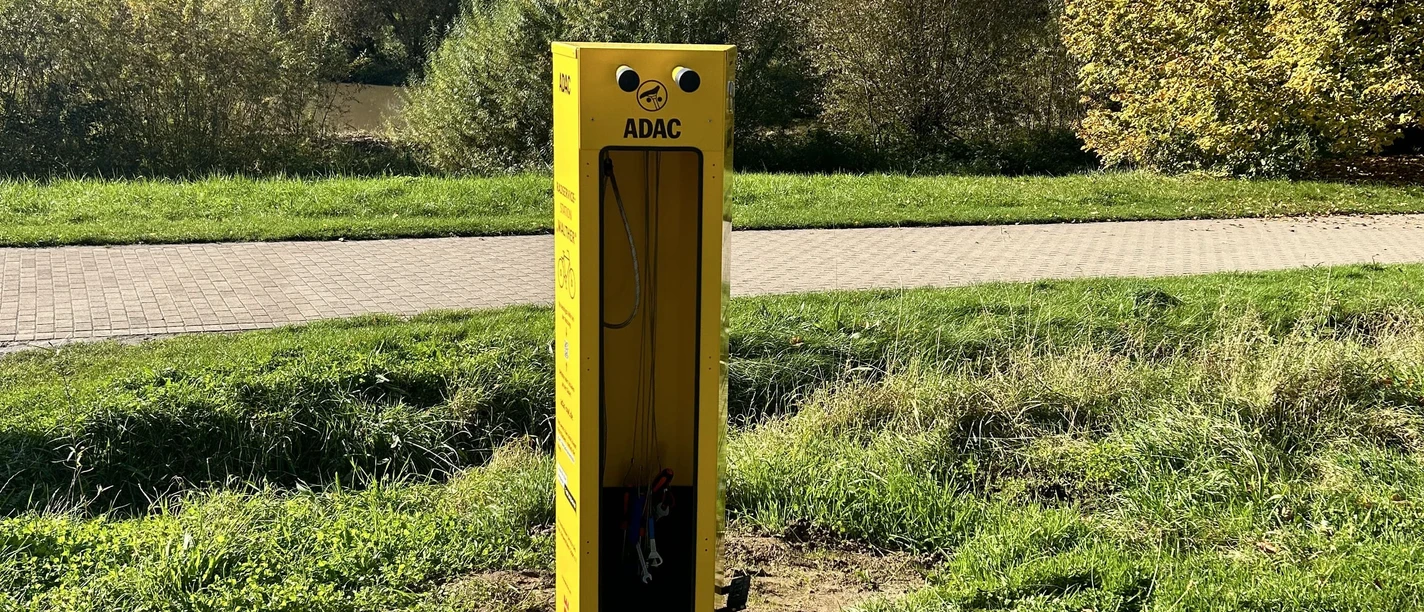 Gelbe ADAC-Fahrradreparatursäule auf Wiese, umgeben von Bäumen und einem Gehweg im Park.
