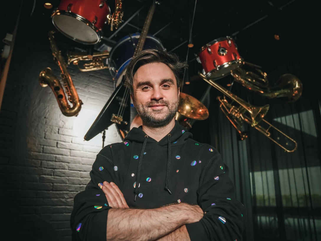 Ein Mann mit Bart steht lächelnd in einem Musikzimmer, umgeben von schwebenden Instrumenten.