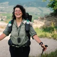 Eine lachende Wanderin mit Rucksack und Stöcken, Landschaft mit Hügeln im Hintergrund.