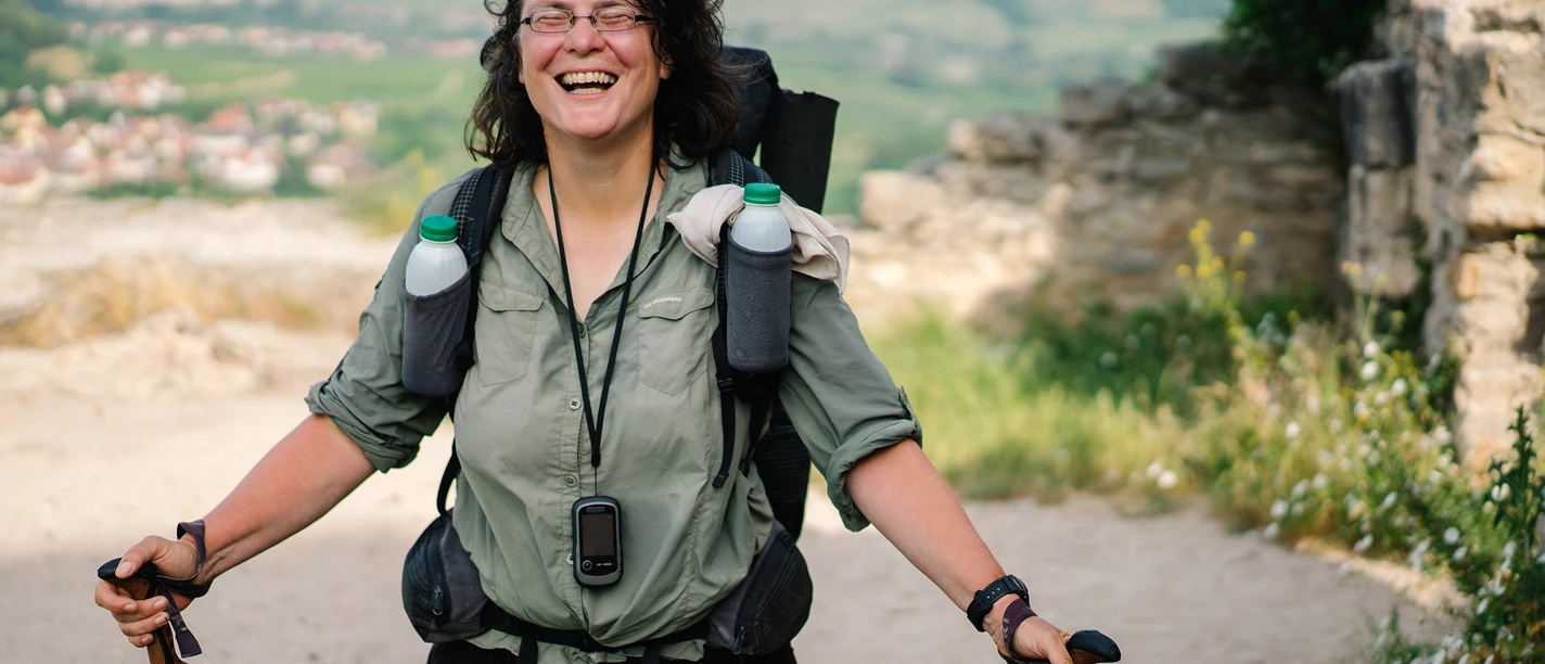 Eine lachende Wanderin mit Rucksack und Stöcken, Landschaft mit Hügeln im Hintergrund.