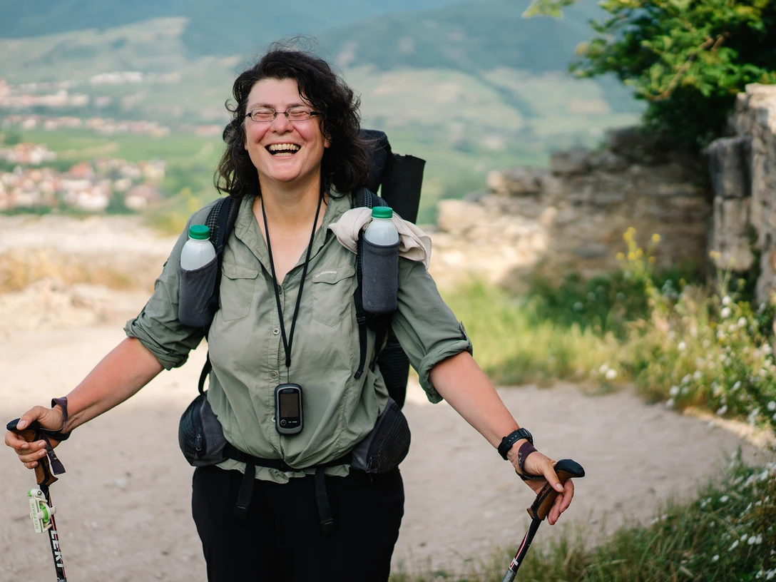 Christine Thürmer Foto Christian Biemann.jpg Eine lachende Wanderin mit Rucksack und Stöcken, Landschaft mit Hügeln im Hintergrund.