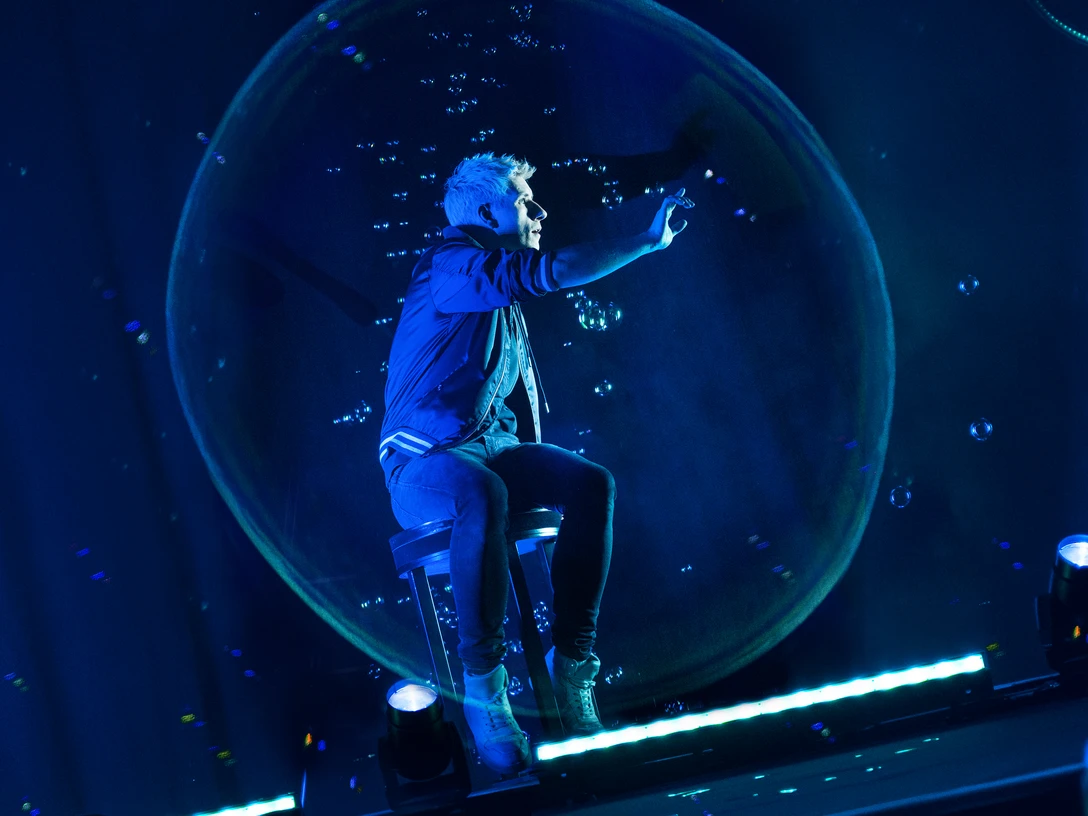 Mellow_Blow-Your-Mind-Tour_Seifenblase(c)mellow.jpg Ein Zauberkünstler in blauer Beleuchtung sitzt auf einem Barhocker und greift nach schwebenden Seifenblasen.