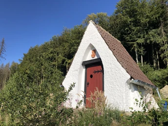 Weiße Lehmkapelle mit roter Tür und steilem Dach, umgeben von Bäumen unter blauem Himmel.