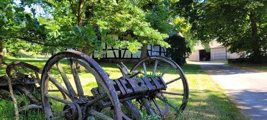 Rossmühle Oberbauerschaft Alte hölzerne Wagenräder liegen unter Bäumen, mit einem Fachwerkhaus und einer Scheune im Hintergrund.