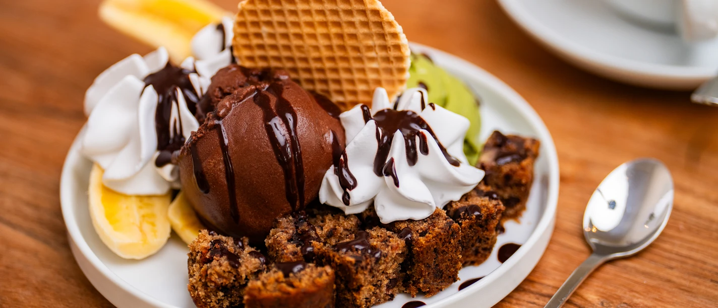 Eiscafé art & wEISe Dekadenter Teller mit Schokoeis, Sahne, knuspriger Waffel, Obst und Kuchenstücken auf Holzplatte.