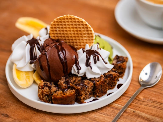 Eiscafé art & wEISe Dekadenter Teller mit Schokoeis, Sahne, knuspriger Waffel, Obst und Kuchenstücken auf Holzplatte.