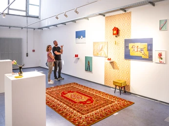 Zwei Personen betrachten Kunstwerke in einer modernen Galerie mit weißer Wand und Teppichboden.