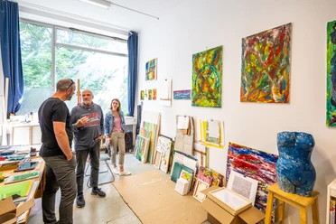 Bielefeld-Ostblock Kulturhaus-Teutoburger-Wald-D-Ketz-230 (1).jpg Drei Personen in einer Künstlerwerkstatt betrachten bunte Gemälde an weißen Wänden mit Fenstern.