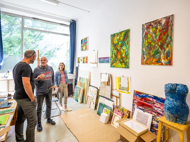 Bielefeld-Ostblock Kulturhaus-Teutoburger-Wald-D-Ketz-230 (1).jpg Drei Personen in einer Künstlerwerkstatt betrachten bunte Gemälde an weißen Wänden mit Fenstern.