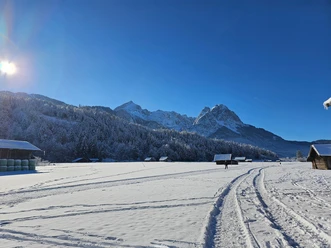 Blick zur Loipe Hausberg - Hammersbach Verschneites Feld mit Loipenspuren, verstreuten Hütten und sonnigem Blick auf winterliche Berge.
