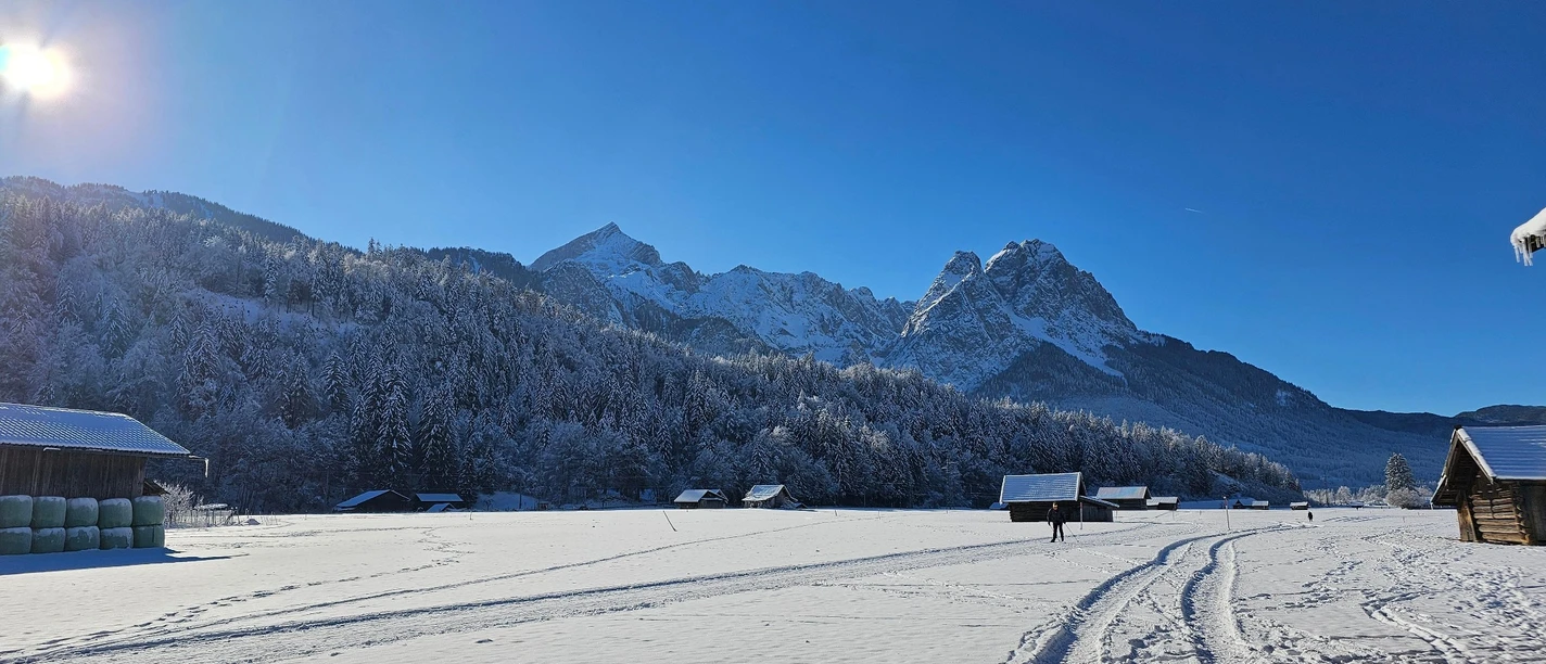 Blick zur Loipe Hausberg - Hammersbach Verschneites Feld mit Loipenspuren, verstreuten Hütten und sonnigem Blick auf winterliche Berge.