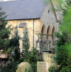 Steinernes Gebäude mit gotischen Fenstern, umgeben von Bäumen und historischen Grabsteinen im Vordergrund.