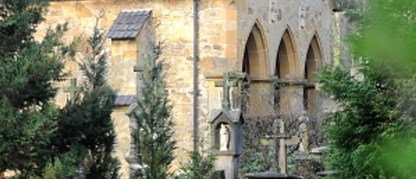 Steinernes Gebäude mit gotischen Fenstern, umgeben von Bäumen und historischen Grabsteinen im Vordergrund.