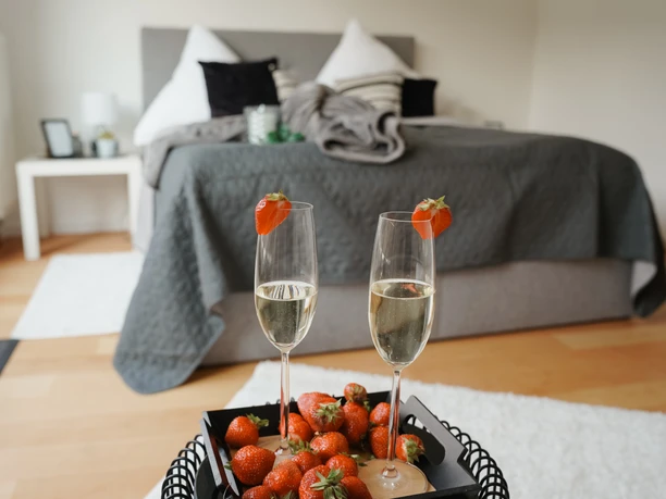 Zwei Sektgläser mit Erdbeeren, umgeben von weiteren Erdbeeren, stehen vor einem Doppelbett.
