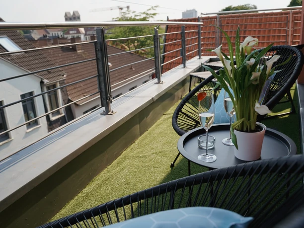 Moderner Balkon mit schwarzen Stühlen, Tisch, Sektgläsern und Blumen, überblickt Stadthäuser.