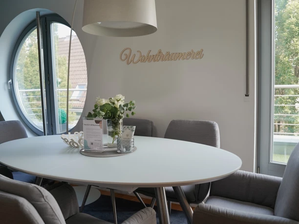 Essgruppe mit ovalem Tisch und sechs grauen Stühlen vor einem Fenster, Blumen als Tischdekoration.