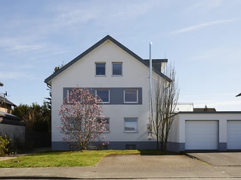 Wohnträumerei - Paderborn Zweistöckiges, weißes Wohnhaus mit blauen Fensterrahmen, Doppelgarage und blühendem Baum vor dem Haus.