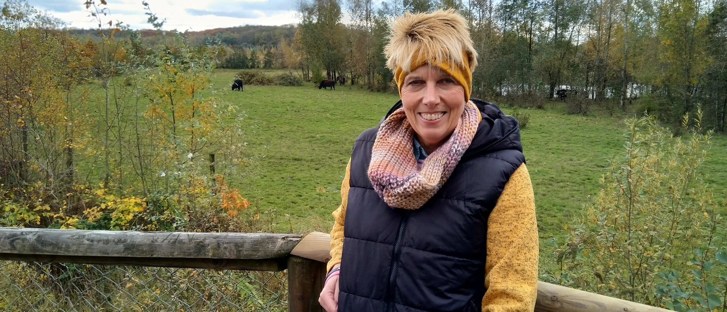Outdoor-Relax Training mit Anja Lindemann Anja Lindemann lächelt in herbstlicher Kleidung an einem Holzzaun vor grüner Wiese mit Bäumen.
