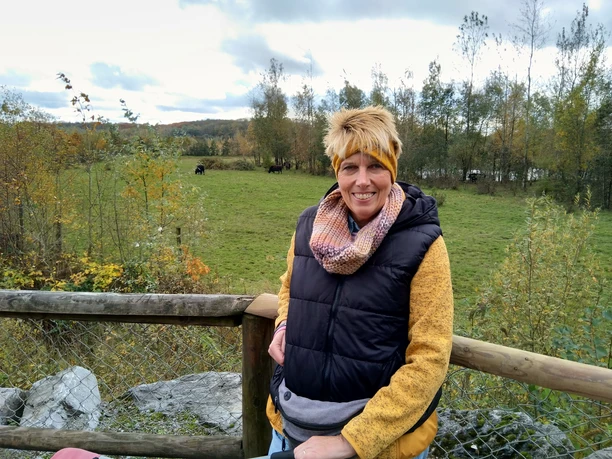 Outdoor-Relax Training mit Anja Lindemann Anja Lindemann lächelt in herbstlicher Kleidung an einem Holzzaun vor grüner Wiese mit Bäumen.