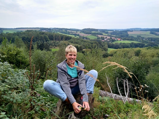 Outdoor-Relax Training mit Anja Lindemann Eine Person sitzt im Freien auf einem Baumstamm, umgeben von grünen Feldern und hügeliger Landschaft.