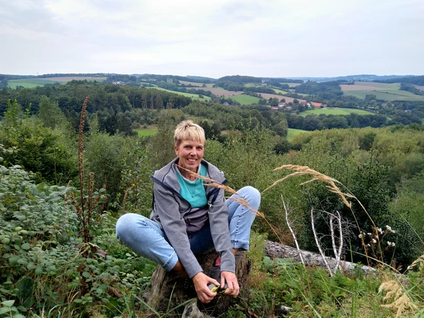 Outdoor-Relax Training mit Anja Lindemann Eine Person sitzt im Freien auf einem Baumstamm, umgeben von grünen Feldern und hügeliger Landschaft.
