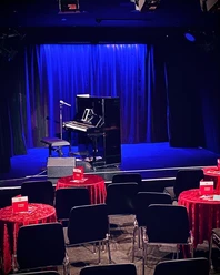 Theater509 / Bürgerhaus Stollwerck Ein gemütlicher Theatersaal mit roten Tischdecken, Stühlen, einem Piano und einem stehenden Mikrofon.