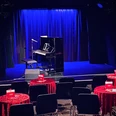 Theater509 / Bürgerhaus Stollwerck Ein gemütlicher Theatersaal mit roten Tischdecken, Stühlen, einem Piano und einem stehenden Mikrofon.A cozy theater hall with red tablecloths, chairs, a piano and a standing microphone.