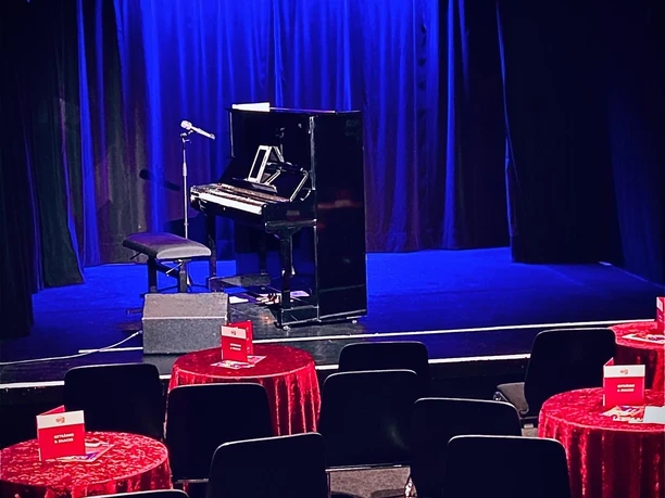 Theater509 / Bürgerhaus Stollwerck A cozy theater hall with red tablecloths, chairs, a piano and a standing microphone.
