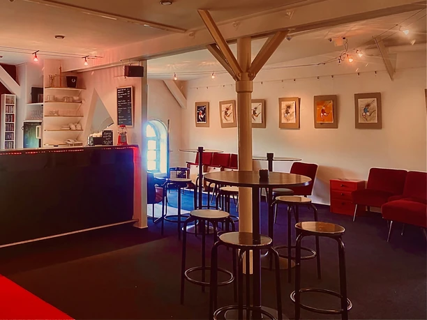 Theater509 / Bürgerhaus Stollwerck Cozy interior of a café with a group of wooden tables, a red couch and a picture gallery on the wall.
