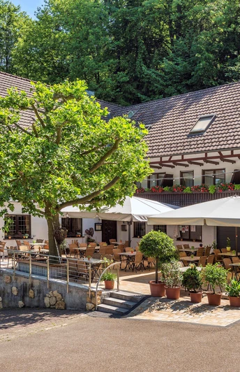 Dreistöckiges, L-förmiges weißes Gebäude mit braunem Dach. Davor Terasse mit hellen Sonnenschirmen und ein Baum