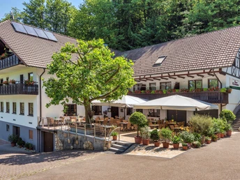 Dreistöckiges, L-förmiges weißes Gebäude mit braunem Dach. Davor Terasse mit hellen Sonnenschirmen und ein Baum