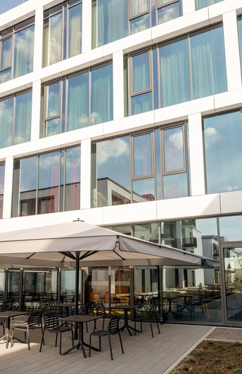 Außenansicht des Hotels mit großer Glasfront. Im Eingangsbereich Stühle und Tische mit Sonnenschirmen