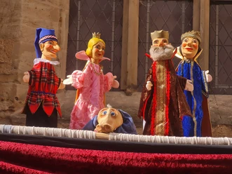 Vijf kleurrijke Hohnstein marionetfiguren in traditionele kleding op een podium voor een stenen gevel.