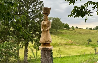 Eine Holzskulptur einer Frau mit Kopfbedeckung steht auf einem Baumstumpf vor einer grünen Wiesenlandschaft.