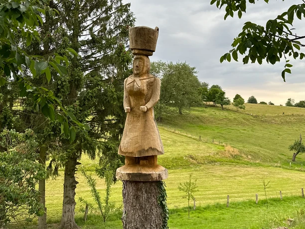 Eine Holzskulptur einer Frau mit Kopfbedeckung steht auf einem Baumstumpf vor einer grünen Wiesenlandschaft.
