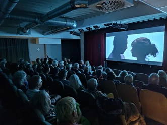 Kino Theaterstuuv.jpg Ein vollbesetztes Kino zeigt eine Projektion von zwei Personen in einem intensiven Profilmoment.