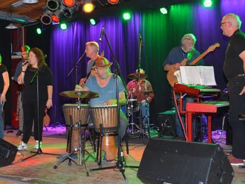 Sechs Musiker performen auf einer Bühne, mehrere Instrumente und bunte Lichter um sie herum.