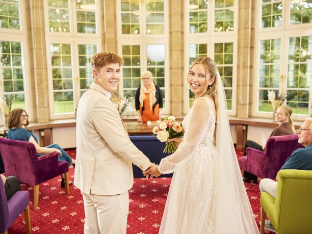 2024_08_30_Herford_Standesamt_Rotunde_0182b.jpg Brautpaar beim Standesamt in Herford in einem hellen Raum mit großen Fenstern und Gästen im Hintergrund.