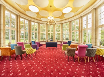 2024_08_30_Herford_Standesamt_Rotunde_0016c.jpg Helle Rotunde mit großen Fenstern, farbenfrohen Stühlen, rotem Teppich und verzierter Decke.