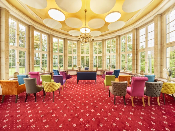 2024_08_30_Herford_Standesamt_Rotunde_0016c.jpg Helle Rotunde mit großen Fenstern, farbenfrohen Stühlen, rotem Teppich und verzierter Decke.