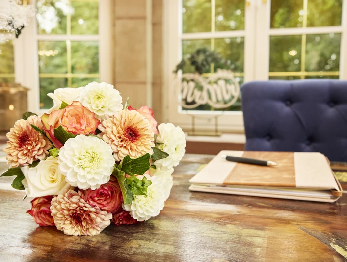 2024_08_30_Herford_Standesamt_Rotunde_0671b.jpg Ein Blumenstrauß in sanften Farben liegt auf einem Holztisch vor einem geöffneten Gästebuch.