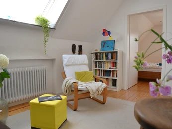 Maries B and B_ Doppelzimmer 2 _M Menke.JPG Helles Zimmer mit Sessel, Bücherregal und Pflanzen; angrenzend an ein Schlafzimmer im Hintergrund.